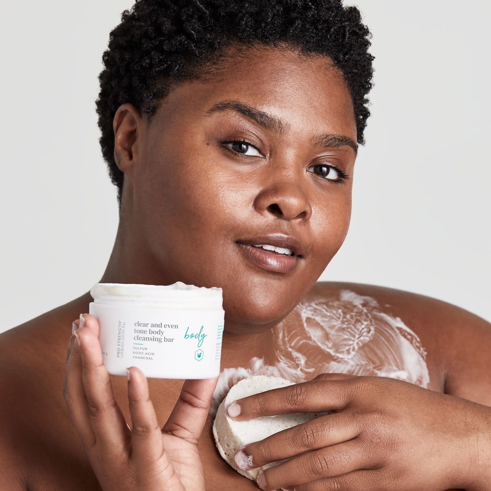 Woman holding jar of Urban Skin RX Clear and Even Tone Body Cleansing Bar and using sponge to apply product on skin
